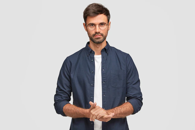 Free Photo | Waist up portrait of handsome serious unshaven male keeps hands together, dressed in dark blue shirt, has talk with interlocutor, stands against white wall. self confident man freelancer