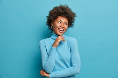 Free Photo | Portrait of dark skinned cheerful woman with curly hair, touches chin gently, laughs happily, enjoys day off, feels happy and enthusiastic, hears something positive, wears casual blue turtleneck